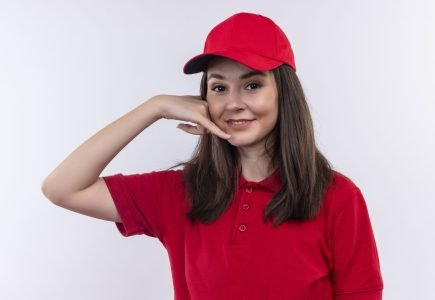 smiling young delivery girl wearing red t-shirt in red cap makes call with her hands on isolated white background